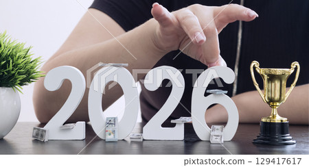 Person pointing at numbers 2026 with cash and trophy, representing financial planning, ambition, and goals for the new year. 129417617