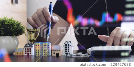 Hand using calculator and pen near miniature houses and stock chart, symbolizing real estate investment planning, valuation, and market strategy. Hand using calculator and pen near miniature houses and stock chart, symbolizing real estate investment planning, valuation, and market strategy. 129417628