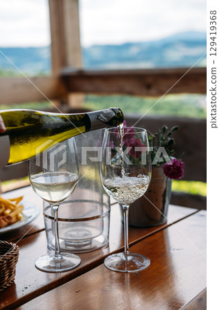 Wine glasses and a bottle sit on a wooden table overlooking a vineyard and rolling countryside. Vineyard dining, wine tasting experience, gourmet rural tourism, slow food Wine glasses and a bottle sit on a wooden table overlooking a vineyard and rolling countryside. Vineyard dining, wine tasting experience, gourmet rural tourism, slow food 129419368