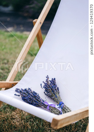 Two small lavender bouquets tied with ribbon rest on a white deck chair in a grassy outdoor setting. Lavender therapy, calming bouquet, self-care plants, holistic wellness. Two small lavender bouquets tied with ribbon rest on a white deck chair in a grassy outdoor setting. Lavender therapy, calming bouquet, self-care plants, holistic wellness. 129419370