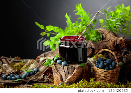 A jar of blueberry jam with fresh berries on an old tree trunk in the forest. A jar of blueberry jam with fresh berries on an old tree trunk in the forest. 129420985