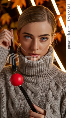 Woman in sweater holding a red ball gag, intense expression with a dramatic background. Woman in sweater holding a red ball gag, intense expression with a dramatic background. 129422181