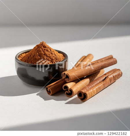 Aromatic Cinnamon Powder and Fresh Sticks in Black Bowl on White Surface Aromatic Cinnamon Powder and Fresh Sticks in Black Bowl on White Surface 129422182