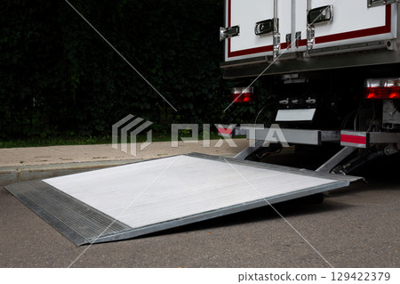 Close-up view of a truck with an unfolded loading ramp on the rear. The ramp is designed for easy loading and unloading of goods. The truck is parked on paved street with green foliage in background 129422379
