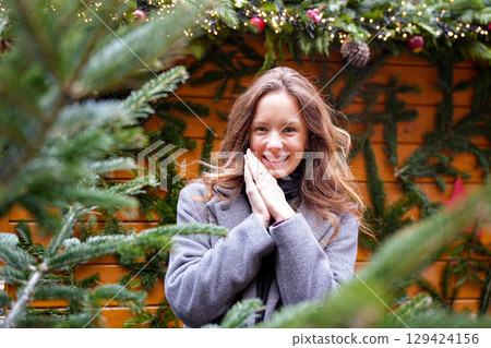 Woman in gray coat smiling near Christmas tree branches outdoors Woman in gray coat smiling near Christmas tree branches outdoors 129424156