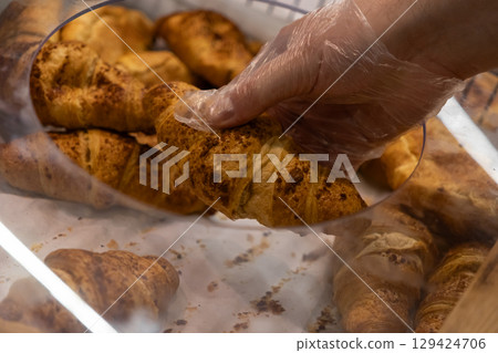 A gloved hand carefully selects a fresh golden croissant from a bakery display, emphasizing hygiene and quality service in food retail A gloved hand carefully selects a fresh golden croissant from a bakery display, emphasizing hygiene and quality service in food retail 129424706