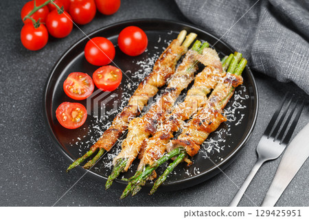 Grilled asparagus wrapped with bacon on black plate with cherry tomatoes on black background 129425951