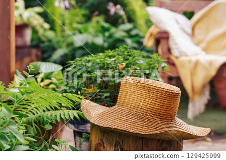 Rustic straw hat resting in lush garden with cozy armchair and soft throw in the background 129425999