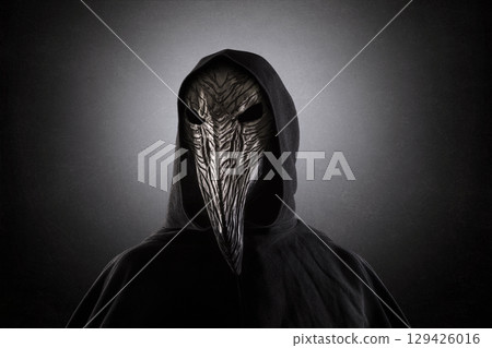 Creepy figure with long bird beak mask in hooded cloak at night over dark misty background 129426016