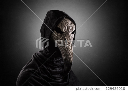Creepy figure with long bird beak mask in hooded cloak at night over dark misty background 129426018