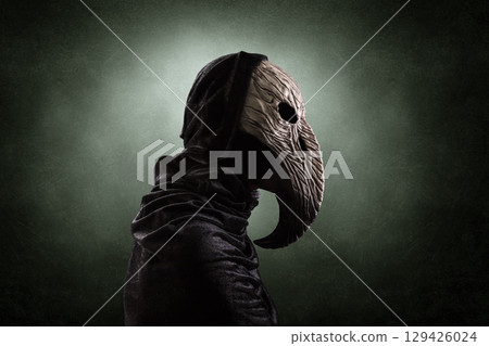 Creepy figure with long bird beak mask in hooded cloak at night over dark misty background 129426024