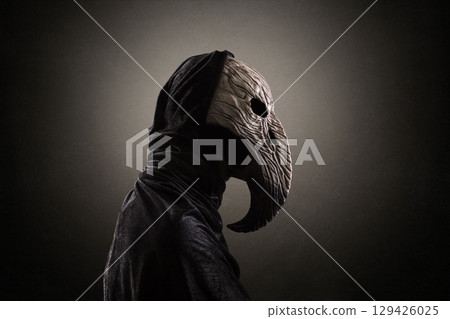Creepy figure with long bird beak mask in hooded cloak at night over dark misty background 129426025