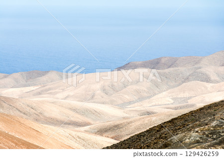 一座荒涼、乾燥、光禿禿的山峰在明媚的陽光下熠熠生輝,遠處的大海清晰可見。富埃特文圖拉島內陸 一座荒涼、乾燥、光禿禿的山峰在明媚的陽光下熠熠生輝,遠處的大海清晰可見。富埃特文圖拉島內陸 129426259