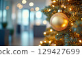 Elegant gold ball on decorated Christmas tree with warm lights in modern office setting during holiday season, blurred background with copy space 129427699