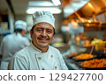 Close-up portrait of full-figured smiling Mexican chef in white uniform clothes with hat on his head, standing in front of working restaurant kitchen and looking at camera 129427700