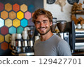 Close-up of portrait of young smiling handsome man, barista in gray t-shirt standing behind counter and with colorful wall of cozy coffee house 129427701