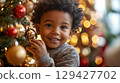 Young smiling african american boy joyfully decorating Christmas tree with colorful ornaments in warmly lit living room during the holiday season 129427702