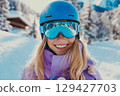 Portrait of smiling young blond girl enjoying sunny day on the slopes with snowy mountain backdrop and ski gear 129427703