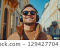 Portrait of happy young caucasian man walking and enjoying music on city street during sunny day, wearing headphones and stylish sunglasses 129427704