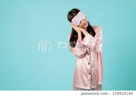Dreamy asian woman sleep with mask cover eye, she is young wearing sleepwear, with a smile, resting her head on her hands, enjoying a blissful night sleep, headshots isolated studio blue background 129428140