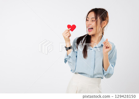 Cheerful asian woman holds red heart, full of love and emotion for valentine day, showing warm smile as lovely, attractive model holds a heart for valentine, headshot isolated studio white background 129428150