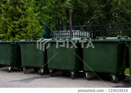 Green trash bins lined up on a city street near a fence, surrounded by trees. The bins are neatly arranged and ready for waste collection, showcasing urban waste management and environmental care Green trash bins lined up on a city street near a fence, surrounded by trees. The bins are neatly arranged and ready for waste collection, showcasing urban waste management and environmental care 129428916