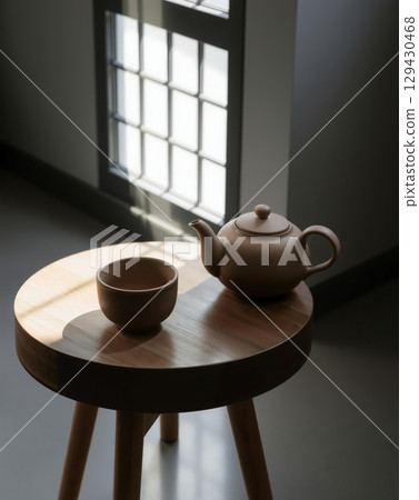 Still life composition featuring a ceramic tea set on a round wooden table near a window. Still life composition featuring a ceramic tea set on a round wooden table near a window. 129430468