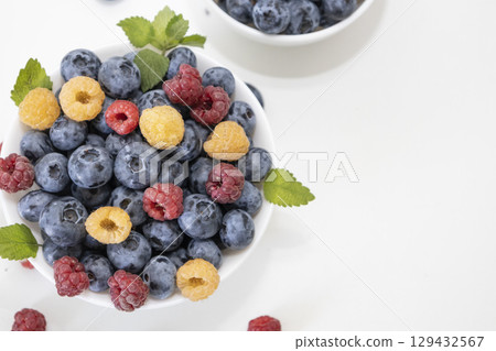 Blueberry and raspberry is tasty, healthy and delicious fruits. Fresh blueberries with red and yellow raspberries with leaves of mint in white bowl. Wild berry in white plate on white background. 129432567