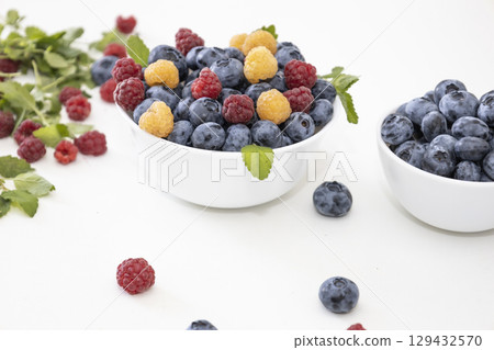 Blueberry and raspberry is tasty, healthy and delicious fruits. Fresh blueberries with red and yellow raspberries with leaves of mint in white bowl. Wild berry in white plate on white background. Blueberry and raspberry is tasty, healthy and delicious fruits. Fresh blueberries with red and yellow raspberries with leaves of mint in white bowl. Wild berry in white plate on white background. 129432570