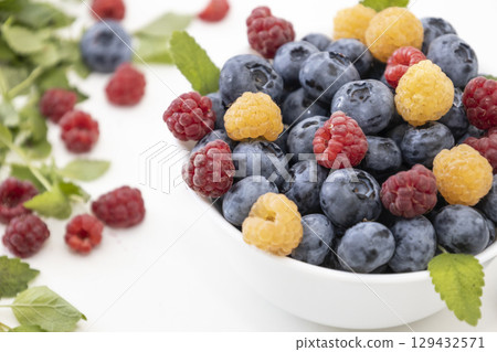 Blueberry and raspberry is tasty, healthy and delicious fruits. Fresh blueberries with red and yellow raspberries with leaves of mint in white bowl. Wild berry in white plate on white background. Blueberry and raspberry is tasty, healthy and delicious fruits. Fresh blueberries with red and yellow raspberries with leaves of mint in white bowl. Wild berry in white plate on white background. 129432571
