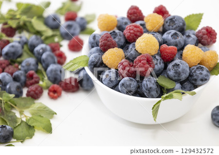 Blueberry and raspberry is tasty, healthy and delicious fruits. Fresh blueberries with red and yellow raspberries with leaves of mint in white bowl. Wild berry in white plate on white background. Blueberry and raspberry is tasty, healthy and delicious fruits. Fresh blueberries with red and yellow raspberries with leaves of mint in white bowl. Wild berry in white plate on white background. 129432572