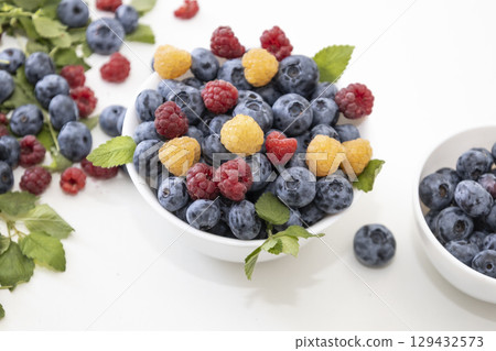 Blueberry and raspberry is tasty, healthy and delicious fruits. Fresh blueberries with red and yellow raspberries with leaves of mint in white bowl. Wild berry in white plate on white background. Blueberry and raspberry is tasty, healthy and delicious fruits. Fresh blueberries with red and yellow raspberries with leaves of mint in white bowl. Wild berry in white plate on white background. 129432573