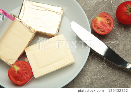 Processed cheese in foil and tomato on wooden plate on grey background Processed cheese in foil and tomato on wooden plate on grey background 129432832