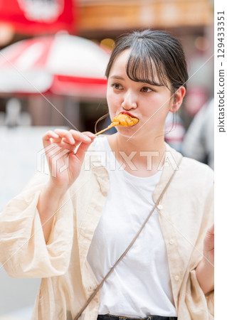 A woman eating kushikatsu (fried skewers) outdoors in Dotonbori, Osaka City, Osaka Prefecture 129433351