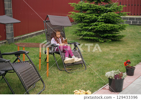 A school-age child is sitting in a chair in the backyard of the house and relaxing while looking at a smartphone. 129433396