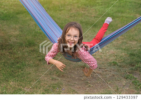 A funny funny child in round glasses is lying on a hammock swinging and laughing in the garden at home. 129433397