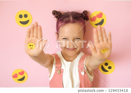 A cute funny girl with pink hair shows emoticons on her palms. A lot of emojis surround the child. World Social Media Day. Smile face yellow 129433418