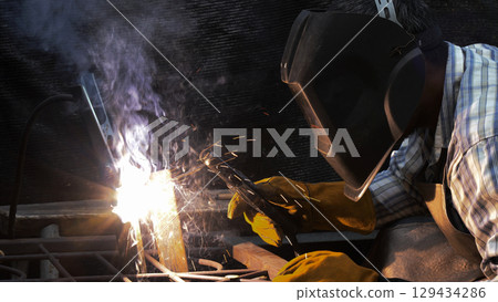 Close-up of focused welder wearing protective helmet and gloves as sparks and thick smoke rise from intense arc welding on metal frame 129434286