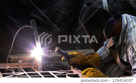 Focused welder wearing helmet and gloves performing arc welding with bright flash on metal surface in workshop Focused welder wearing helmet and gloves performing arc welding with bright flash on metal surface in workshop 129434287