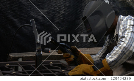 Welding technician in striped shirt and safety gear examining torch position before striking arc in low light workspace Welding technician in striped shirt and safety gear examining torch position before striking arc in low light workspace 129434289