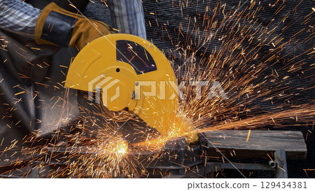 Yellow electric chop saw cutting through metal tube with explosion of bright orange sparks, gloved hands guiding tool during active industrial process in outdoor workshop 129434381