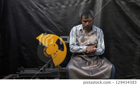 Male factory worker in protective apron sitting beside industrial cutting saw, examining hand with tired expression in manufacturing work environment Male factory worker in protective apron sitting beside industrial cutting saw, examining hand with tired expression in manufacturing work environment 129434383