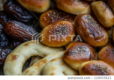 A rustic wooden plate holds a delicious arrangement of glossy dates and golden-brown pastries. The scene is illuminated by soft, crescent moon-shaped lights. 129435621