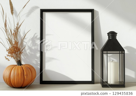 A blank, vertical black picture frame is a centerpiece surrounded by autumn decor. The decorations include pumpkins, lanterns with candles, dried leaves, and tall grasses. 129435836