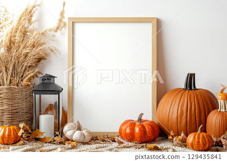 A blank, vertical black picture frame is a centerpiece surrounded by autumn decor. The decorations include pumpkins, lanterns with candles, dried leaves, and tall grasses. 129435841