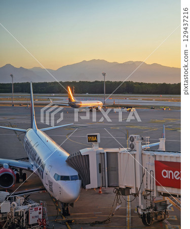 ANTALYA, TURKEY -  JULY 5, 2025 Antalya airport scene at sunset and a plane docked at the jet bridge with other aircrafts visible on the tarmac. Distant mountains range silhouette in the background 129437216