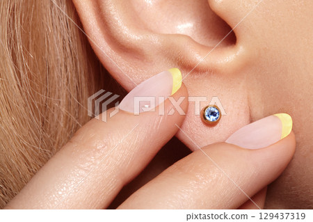 Close-up macro shot of female ear with gold stud earring featuring round blue gemstone. Elegant minimalist jewelry detail with polished metal and sparkling stone 129437319