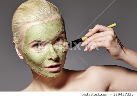 Beautiful Woman Applying Green Facial Mask. Beauty Treatments. Spa Girl Apply Clay Facial mask on grey background Beautiful Woman Applying Green Facial Mask. Beauty Treatments. Spa Girl Apply Clay Facial mask on grey background 129437322