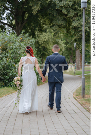 Bride and groom walking away hand in hand down a curved park pathway, surrounded by lush green trees. The bride holds a cascading bouquet of roses 129437336