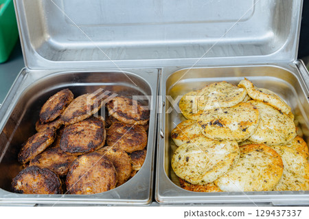 Close up of stainless buffet trays filled with grilled patties and herb chicken patties. Useful for catering companies, menu photography, hospitality marketing and event planning visuals. 129437337